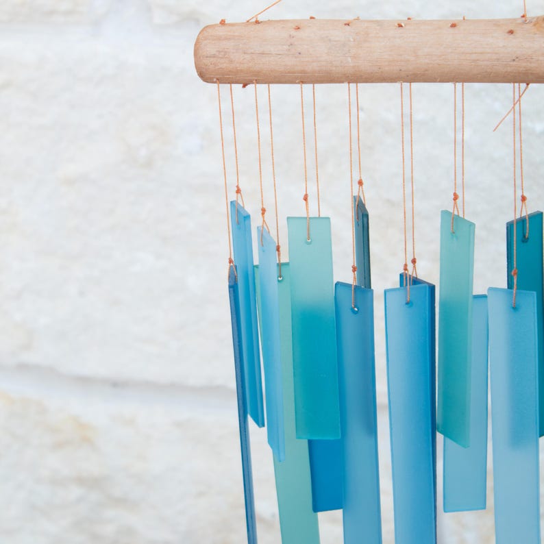 May include: A wind chime featuring multiple rectangular frosted glass pieces in various shades of blue and teal. The glass rectangles are suspended by thin orange strings from a horizontal piece of light brown natural driftwood.