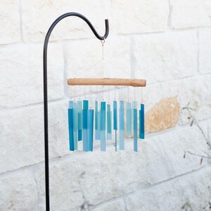 May include: A wind chime featuring multiple rectangular glass pieces in varying shades of blue and teal, suspended by thin wires from a horizontal piece of light-colored driftwood. The chime hangs from a black metal shepherd's hook stand.