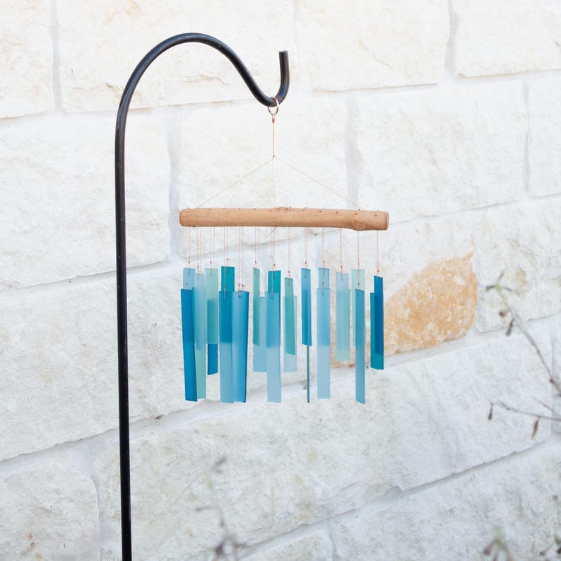 May include: A wind chime featuring multiple rectangular glass pieces in varying shades of blue and teal, suspended by thin wires from a horizontal piece of light-colored driftwood. The chime hangs from a black metal shepherd's hook stand.