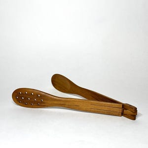 May include: Wooden kitchen tongs with a natural finish. One side features a series of small holes, while the other has a solid, curved design. The tongs are designed with a folding mechanism.