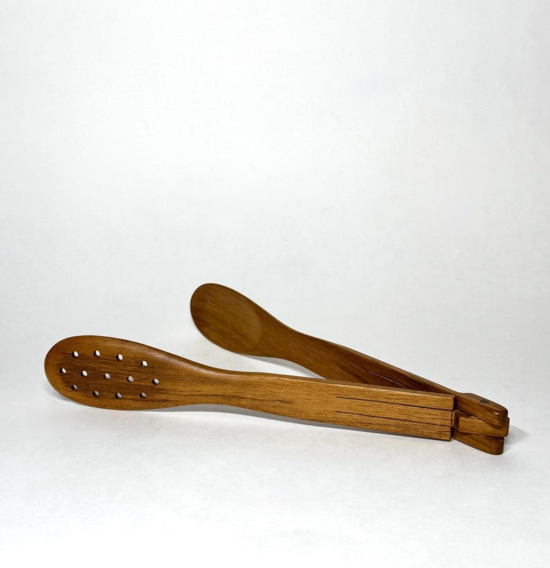 May include: Wooden kitchen tongs with a natural finish. One side features a series of small holes, while the other has a solid, curved design. The tongs are designed with a folding mechanism.