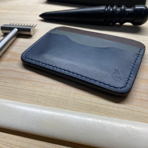 Puede incluir: Una cartera de cuero azul marino con un borde cosido y un pequeño logotipo. La cartera tiene tres ranuras para tarjetas y un bolsillo más grande para efectivo.