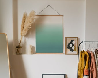 Op de afbeelding: Interieur van een kamer met ingelijste kunstwerken en decoratieve elementen. Een grote print met een groen en blauw verloop hangt boven een kleine tafel met een ingelijste zwart-witfoto. Een kledingrek met diverse kledingstukken is zichtbaar.