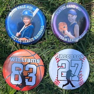 Puede incluir: Cuatro botones redondos con temas deportivos. Uno presenta a un jugador de b&eacute;isbol con el texto "SHAON VALE #22 LITTLE LEAGUE BASEBALL". Otro muestra una pelota de baloncesto con "WILLIAMS 83". El tercero tiene un jugador de b&eacute;isbol con "JACOBSON 27". El &uacute;ltimo tiene una ni&ntilde;a con "PLAYER NAME #00 TEAM LEAGUE NAME".