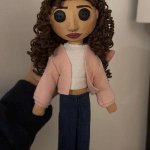 May include: Handmade doll with button eyes, brown curly hair, and a painted face. The doll wears a pink jacket, white top, and blue jeans. A unique, handcrafted art doll.