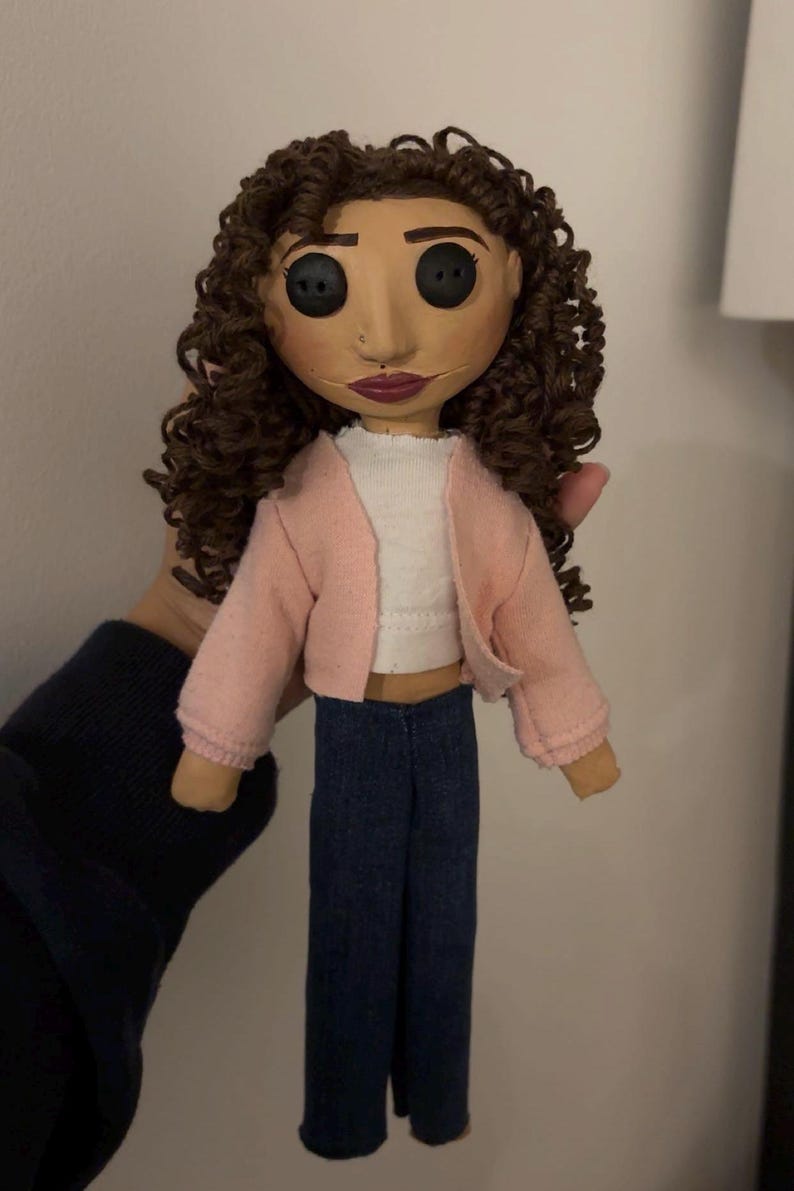 May include: Handmade doll with button eyes, brown curly hair, and a painted face. The doll wears a pink jacket, white top, and blue jeans. A unique, handcrafted art doll.