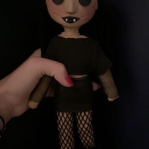 May include: A handmade doll with button eyes, black hair, and a stitched face. The doll wears a black crop top, skirt, and fishnet stockings. The doll's mouth is open, revealing small fangs. The doll is held against a dark background.