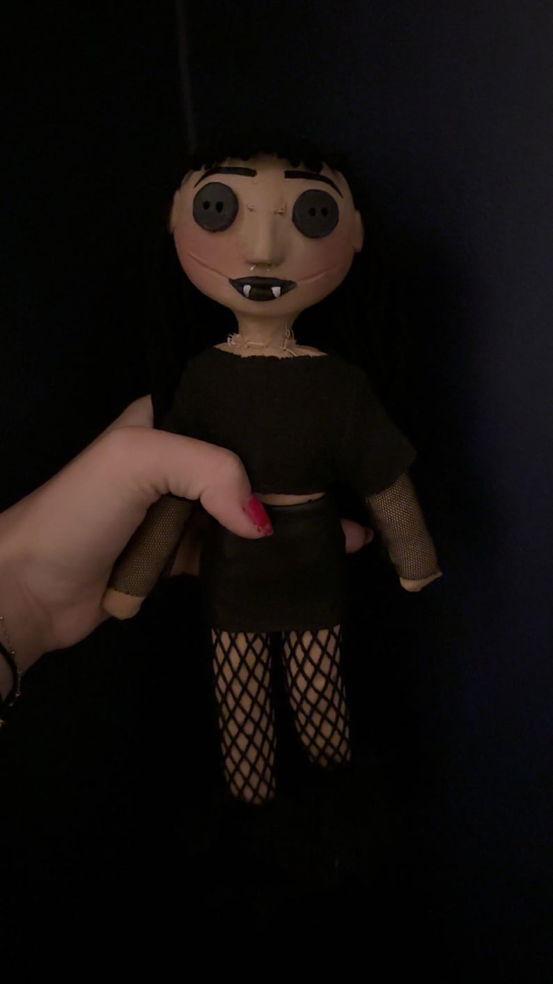 May include: A handmade doll with button eyes, black hair, and a stitched face. The doll wears a black crop top, skirt, and fishnet stockings. The doll's mouth is open, revealing small fangs. The doll is held against a dark background.