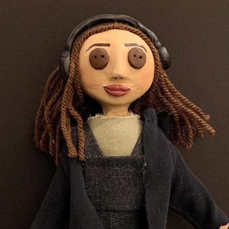 May include: A handmade doll with button eyes, brown braided hair, and a painted face. The doll wears a dark blue jacket over a gray dress and black headphones. The doll is a unique, handcrafted item.