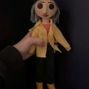 May include: A handmade doll with button eyes, gray hair, and a painted face. The doll wears a yellow jacket over a red shirt, black pants, and yellow shoes. The doll is a character from the animated film "Coraline."