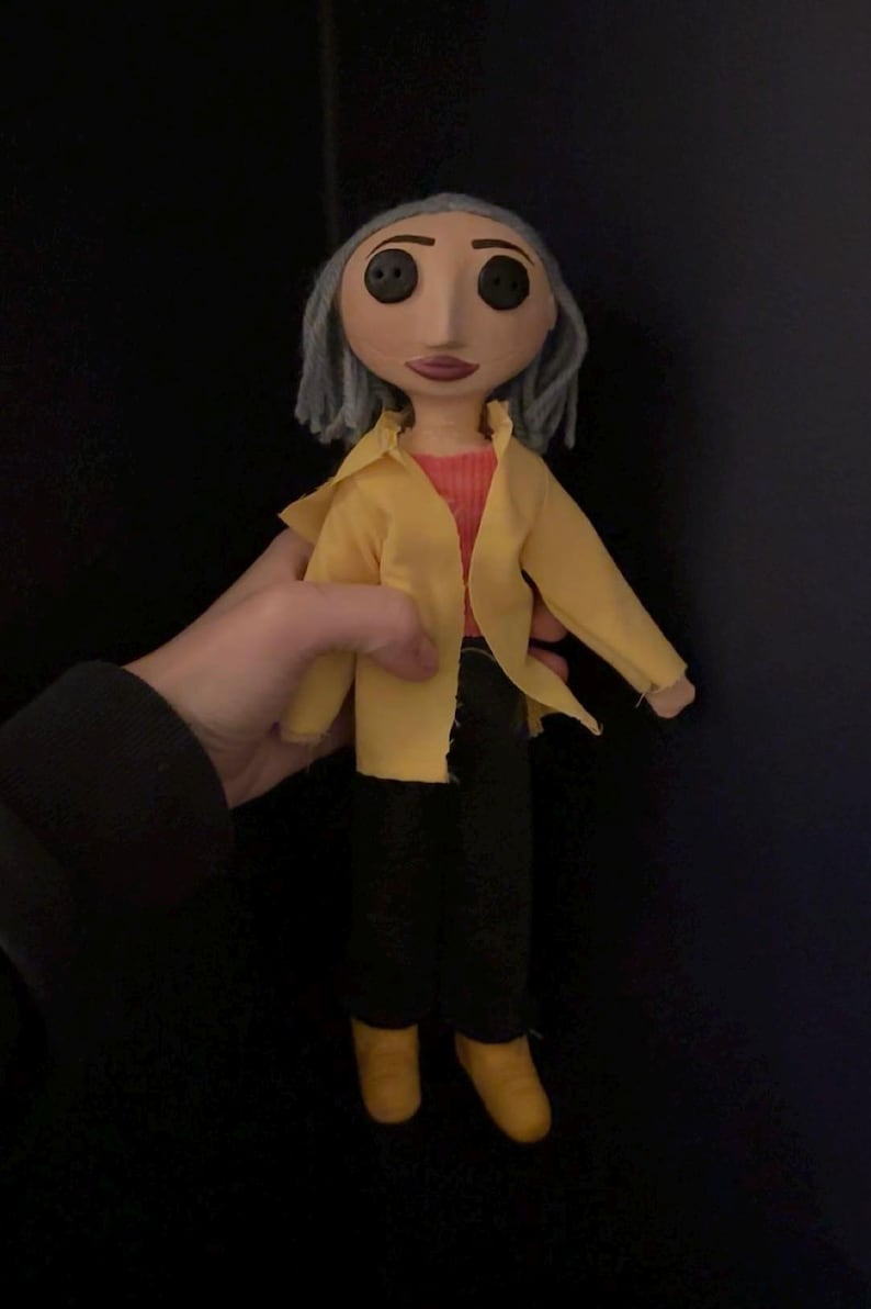 May include: A handmade doll with button eyes, gray hair, and a painted face. The doll wears a yellow jacket over a red shirt, black pants, and yellow shoes. The doll is a character from the animated film "Coraline."