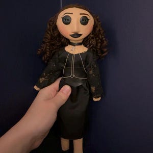 May include: A handmade doll with a fabric face, button eyes, and curly brown hair. The doll wears a black dress with lace sleeves and a silver chain necklace. The doll is held against a dark blue background.