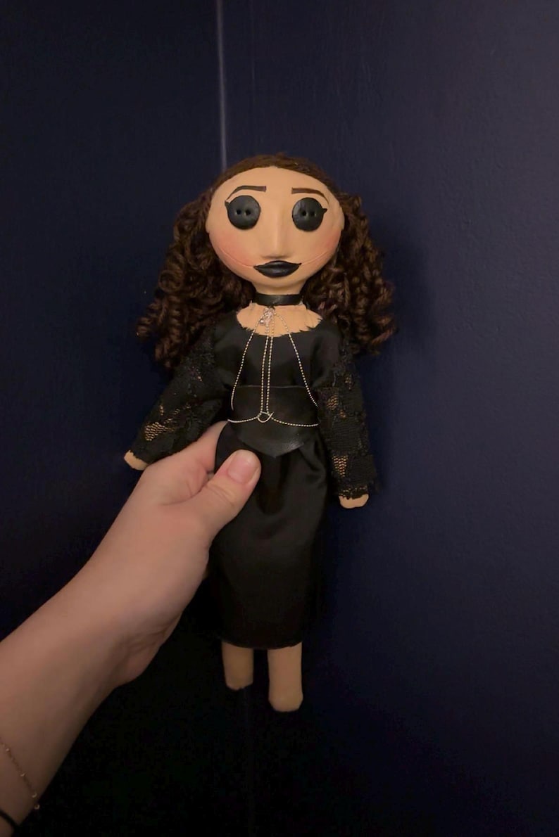 May include: A handmade doll with a fabric face, button eyes, and curly brown hair. The doll wears a black dress with lace sleeves and a silver chain necklace. The doll is held against a dark blue background.