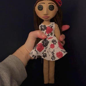 May include: A handmade doll with button eyes, brown hair, and a red bow. The doll wears a white dress with a red and black floral pattern. The doll's legs are tan, and the doll is held by a person.