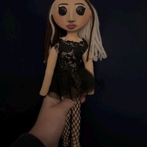 May include: A handmade doll with a fabric face, black button eyes, and a black lace dress. The doll has brown and white yarn hair and is wearing fishnet stockings. The doll is held against a dark blue background.
