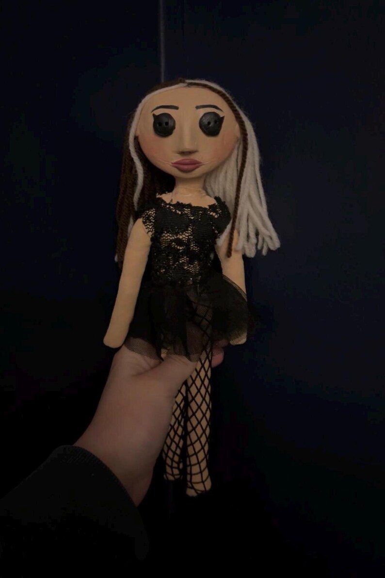 May include: A handmade doll with a fabric face, black button eyes, and a black lace dress. The doll has brown and white yarn hair and is wearing fishnet stockings. The doll is held against a dark blue background.