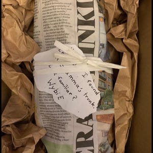 May include: A package wrapped in newspaper and brown paper, inside a cardboard box. A handwritten note on the package reads "I found emma's trunk. Wybi E familiar?"