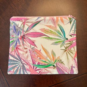 May include: A rectangular zippered pouch featuring an off-white background printed with a repeating pattern of cannabis leaves in vibrant shades of pink, purple, orange, green, and blue. The pouch has a light pink zipper closure along the top edge.