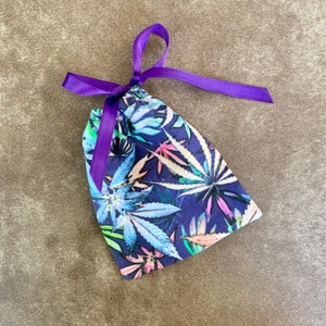 May include: A small, drawstring bag with a vibrant botanical print featuring cannabis leaves in shades of blue, green, and pink against a dark blue background. The bag is tied with a purple satin ribbon.