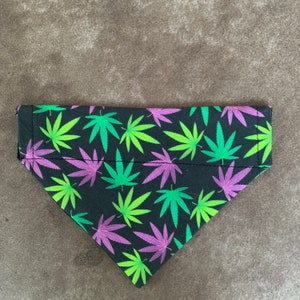 May include: A black pet bandana with a repeating pattern of green and purple cannabis leaves. The bandana is triangular in shape and has a folded edge.