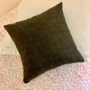 May include: A dark brown square pillow with a repeating pattern of colorful, abstract shapes. The pillow is resting on a white bedspread with a quilted diamond pattern and a white pillow with a red floral design.