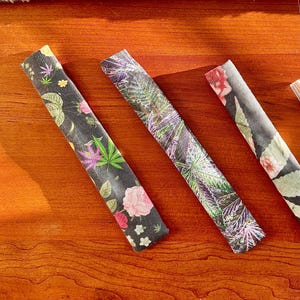 May include: Three long, narrow, rectangular fabric strips with distinct patterns. The left strip features a black background with pink and green cannabis leaves and pink flowers. The middle strip has a dark, leafy botanical print. The right strip shows a floral pattern.