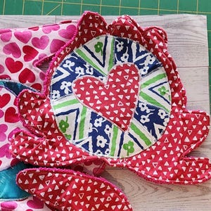 May include: A fabric potholder featuring a central circular design with a red heart surrounded by blue and green floral and geometric patterns. The outer edge is scalloped and red with white triangles, with additional heart-patterned fabric visible.