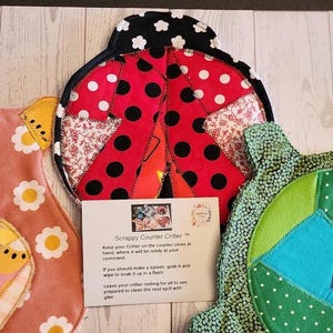 May include: Three fabric kitchen cloths shaped like animals, including a red ladybug with black polka dots, a pink cloth with white flowers, and a green cloth with geometric patterns. A white card with the text Scrappy Counter Critter is included.