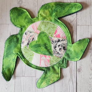 May include: A handmade, green fabric turtle with a patchwork design on its shell. The turtle's body is a vibrant green, and the shell features a circular patchwork of floral and geometric patterns in pink, white, and gray.