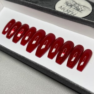 May include: A set of ten press-on nails in a box. The nails are a deep red colour, with a glossy finish. Some nails have red glitter accents. The nails are rectangular in shape.