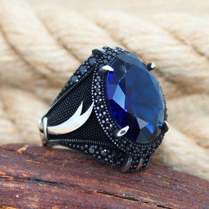 May include: A silver ring with a large, oval, deep blue gemstone as the centerpiece. The ring has a black band with intricate detailing and smaller black stones around the main gem. A crescent moon design is visible on the side.