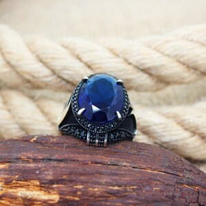 May include: A silver ring with a large, oval, deep blue gemstone as the centerpiece. The ring is set with smaller black stones around the main stone and on the band. The ring is displayed on a piece of wood.