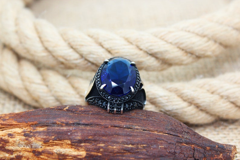 May include: A silver ring with a large, oval, deep blue gemstone as the centerpiece. The ring is set with smaller black stones around the main stone and on the band. The ring is displayed on a piece of wood.