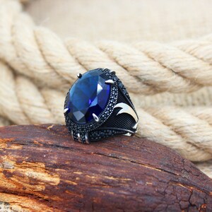 May include: A silver ring with a large, oval, deep blue gemstone as the centerpiece. The ring is set with smaller black stones around the main gem and on the band. The ring rests on a piece of wood, with a rope in the background.