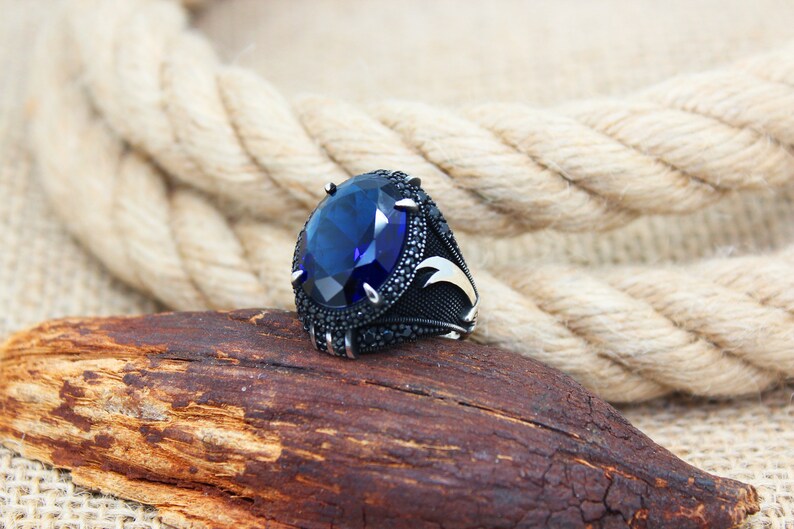 May include: A silver ring with a large, oval, deep blue gemstone as the centerpiece. The ring is set with smaller black stones around the main gem and on the band. The ring rests on a piece of wood, with a rope in the background.