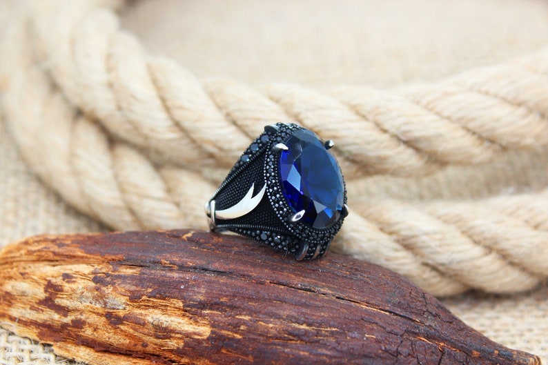 May include: A silver ring with a large, oval, deep blue gemstone as the centerpiece. The ring has a black band with intricate detailing and smaller black stones around the main gem. A crescent moon design is visible on the side.