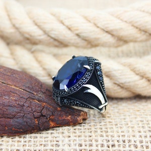 May include: A silver ring with a large, oval-cut, deep blue gemstone as the centerpiece. The ring's band is black with intricate detailing and smaller black stones. The design includes a stylized, curved accent on the sides.