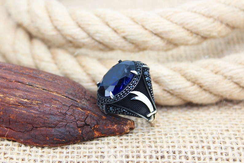 May include: A silver ring with a large, oval-cut, deep blue gemstone as the centerpiece. The ring's band is black with intricate detailing and smaller black stones. The design includes a stylized, curved accent on the sides.