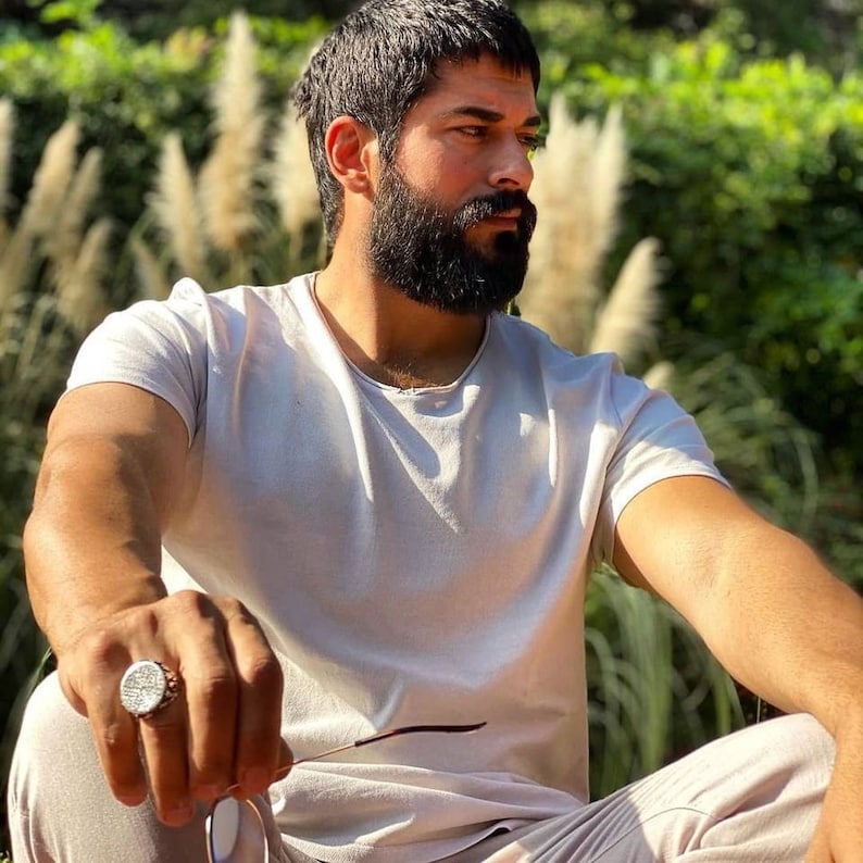 Puede incluir: Un hombre con barba y pelo corto y oscuro lleva una camiseta blanca y pantalones beige. Est&aacute; sentado al aire libre con unas gafas de sol en la mano. Lleva un anillo de plata en la mano izquierda.