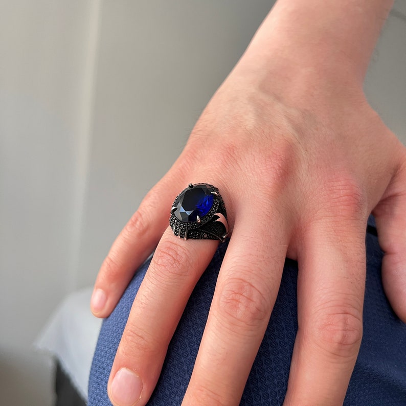 May include: A black ring with a large, oval-shaped, deep blue gemstone. The ring has a detailed design with smaller black stones around the main stone and on the band. A silver accent is visible on the side of the ring.