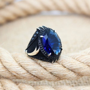 May include: A silver ring with a large, oval-shaped, deep blue gemstone. The ring has a black band with small black stones and gold accents. The ring is set against a background of a thick, woven rope.