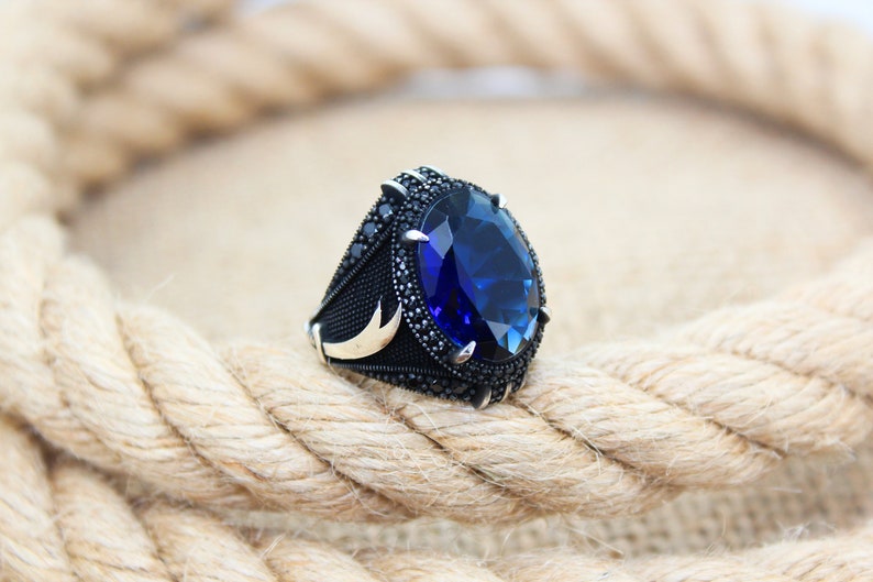 May include: A silver ring with a large, oval-shaped, deep blue gemstone. The ring has a black band with small black stones and gold accents. The ring is set against a background of a thick, woven rope.