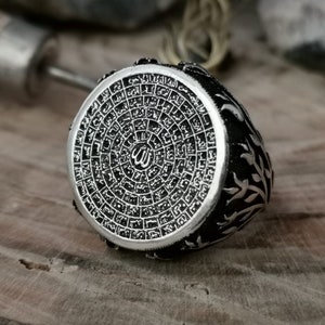 Puede incluir: Un anillo de plata con un dise&ntilde;o intrincado que presenta escritura &aacute;rabe y un s&iacute;mbolo isl&aacute;mico estilizado. El anillo est&aacute; sobre un fondo de madera.