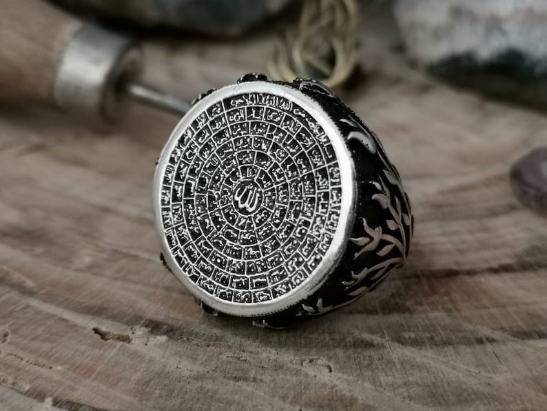 Puede incluir: Un anillo de plata con un dise&ntilde;o intrincado que presenta escritura &aacute;rabe y un s&iacute;mbolo isl&aacute;mico estilizado. El anillo est&aacute; sobre un fondo de madera.