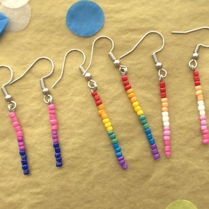 May include: Six pairs of rainbow-coloured beaded earrings with silver hooks. The earrings are arranged in a row on a brown surface.