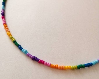 May include: A beaded necklace featuring a rainbow of colors. The necklace is made of small, round beads strung together, transitioning from red to orange, yellow, green, blue, and purple. The necklace has a silver clasp.
