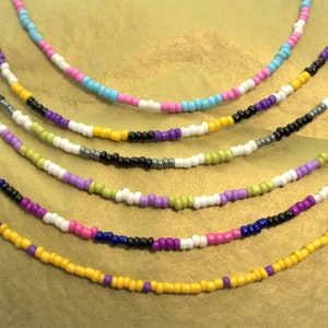 Op de afbeelding: Een set van zeven kralenkettingen in verschillende kleuren, waaronder geel, paars, roze, blauw, wit, groen en zwart. De kettingen zijn in een gelaagd patroon op een gouden achtergrond gerangschikt.