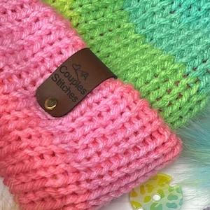 May include: A pink, green, and yellow striped crocheted headband with a brown leather tag that says "Couples Stitches".