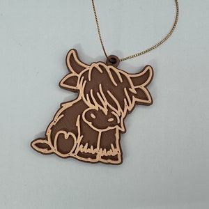 Puede incluir: Un adorno de madera marrón con forma de vaca de las tierras altas con una cuerda dorada para colgar.