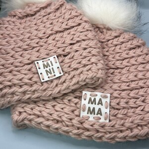 May include: Two pink knitted hats with white pom poms. The smaller hat has a white label that says "MINI" and the larger hat has a white label that says "MAMA".